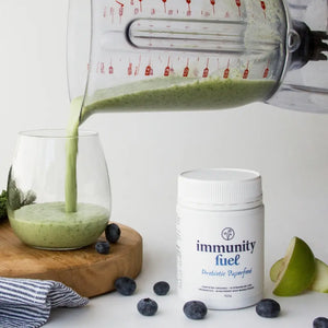Green smoothie being poured into a glass with 'immunity fuel' supplement container on a white background