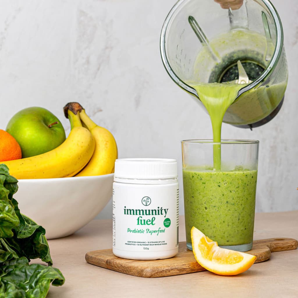 Green smoothie being poured into a glass with immunity fuel powder for bloating, fruits, and vegetables on a table.