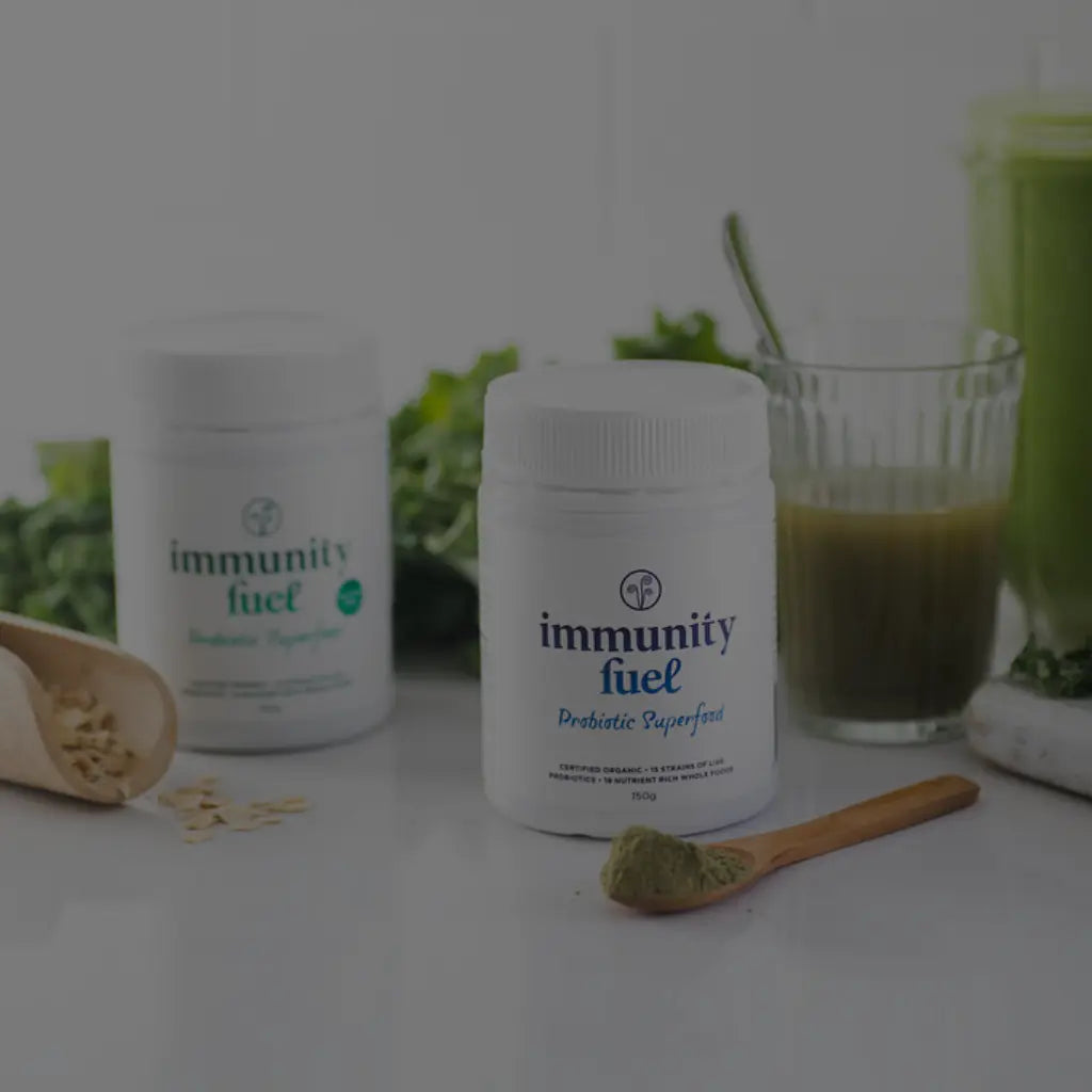 Two supplement jars labeled 'immunity fuel' on a white surface, with a glass of green drink, a wooden spoon of powder, and green herbs in the background. Best probiotics NZ