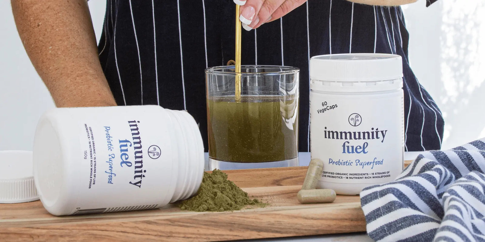 Person pouring a green powder into a glass with 'immunity fuel' supplement containers on a wooden surface.