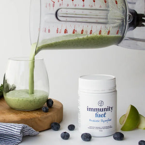 Green smoothie being poured into a glass with 'immunity fuel' supplement and blueberries on a white background
