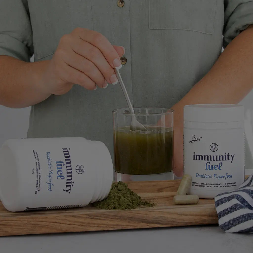 A person in a gray button-up shirt stirs a green drink with a spoon, with two white tubs of 'immunity fuel' probiotic superfood and a small pile of green powder on a wooden board in the foreground.