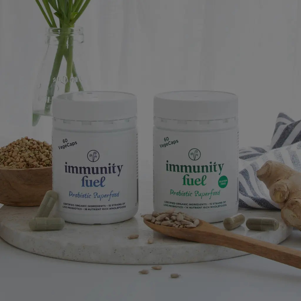 Two white supplement jars labeled 'immunity fuel' with capsules and ingredients arranged on a marble board with ginger, a wooden spoon, and a glass vase in the background.