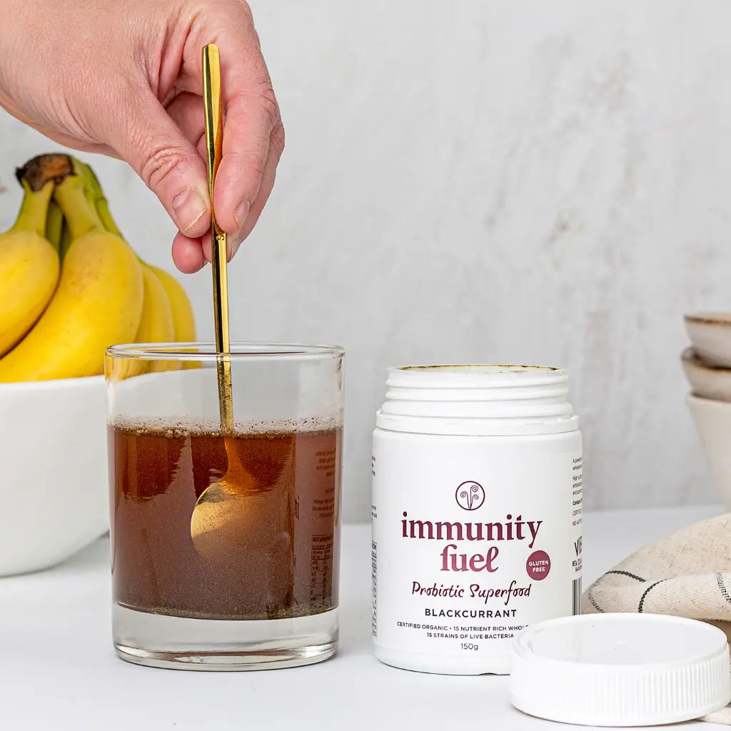Glass of liquid with a spoon, jar labeled 'immunity fuel', and bananas on a light surface.
